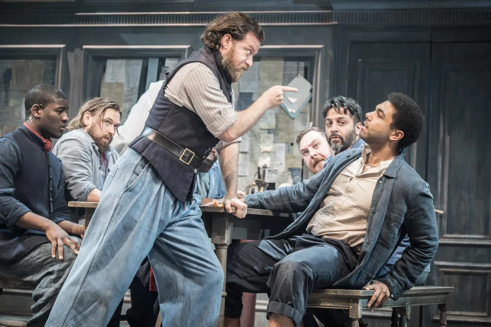 A man wearing a black waistcoat and blue trouser stands in the centre of the image holding a notebook toward another man sat at a table.