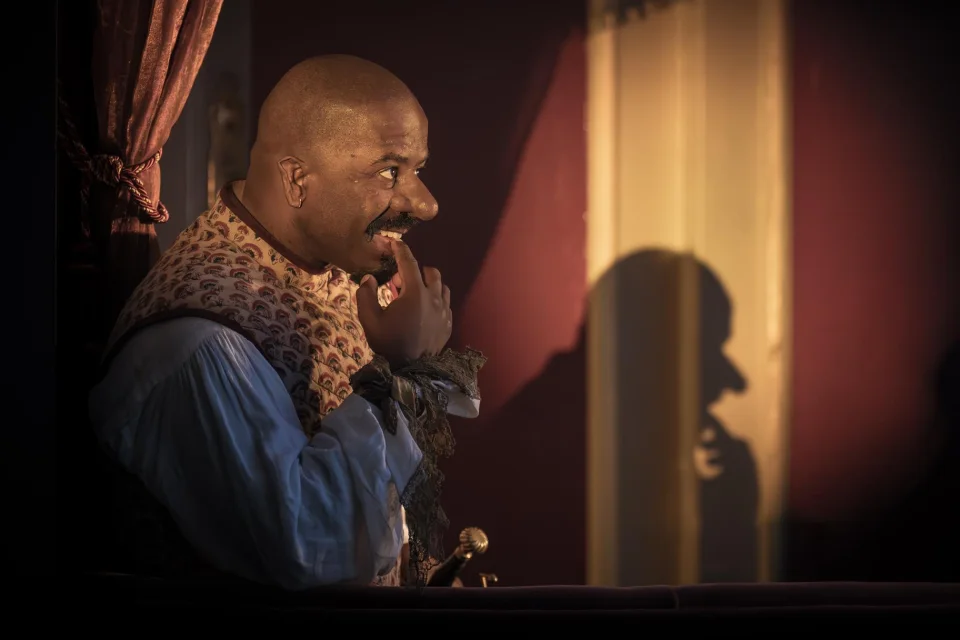 A man wearing a large prosthetic nose is in the centre of the image, he wears a patterned waistcoat and blue shirt and holds a finger on his lips. An outline of his shadow can be seen on the wall behind.