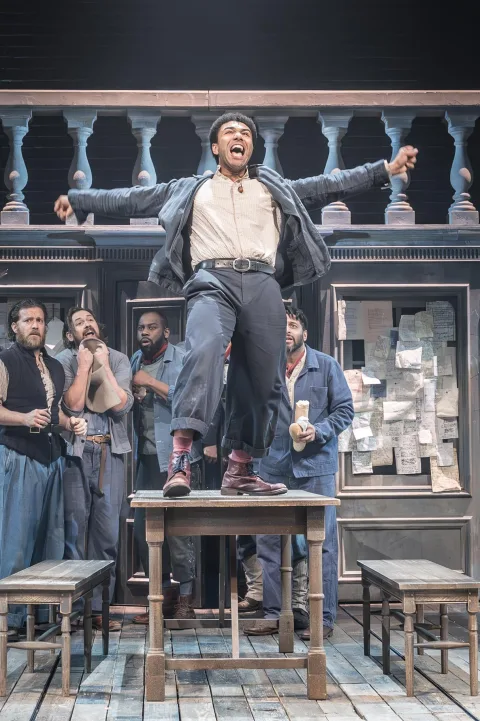A young man stands on a large wooden table shouting and smiling. A group of young men stand behind him watching.