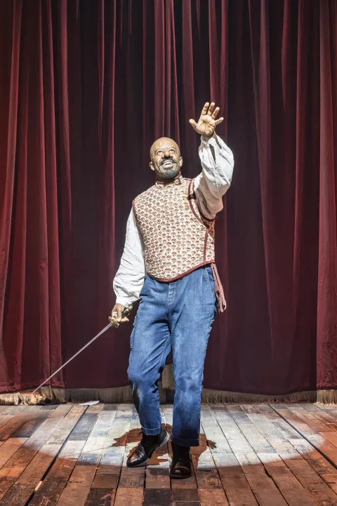 A man wearing a large prosthetic nose stands in the centre of the image with a sword in his left hand and his right hand raised upwards. He delivers a speech.