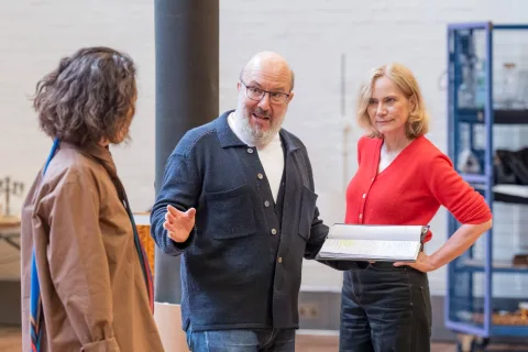 Three adults stand indoors talking; a man with glasses holds a folder and gestures, while two women listen, one wearing a red jumper and the other a brown jacket.