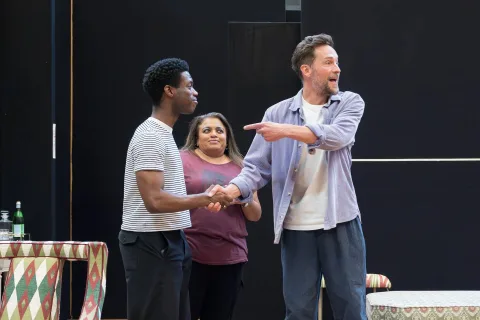 Three people in a rehearsal room; two men shake hands whilst one gestures and speaks, and a woman stands behind them smiling. A table with a patterned cloth and a water bottle is nearby.
