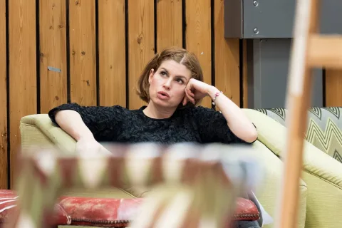 A woman with short brown hair sits on a sofa, resting her head on her hand and looking up with a thoughtful expression. Wooden panelled wall is in the background.