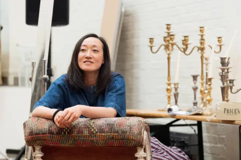 A woman with long dark hair sits on a patterned chair, leaning forward and smiling; behind her are gold candlesticks and a Price’s candle box on a wooden table.
