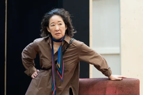 A woman with curly hair wearing a brown shirt and a colourful scarf leans on a red surface, looking to the side with a thoughtful expression.