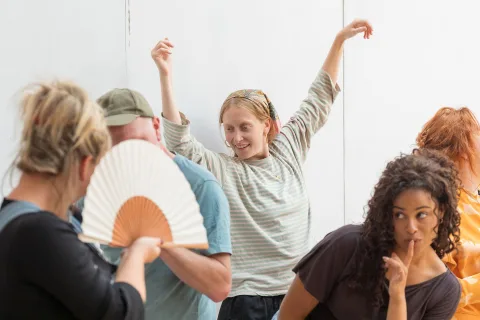 A group of people move in various ways, in the centre stands a white, tall, ginger woman with freckles who wears a bandana and a long-sleeved striped t-shirt, with her arms in the air above her head.