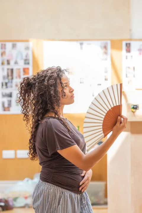 An average height, mixed race girl stands and holds an open fan in a brightly lit room.