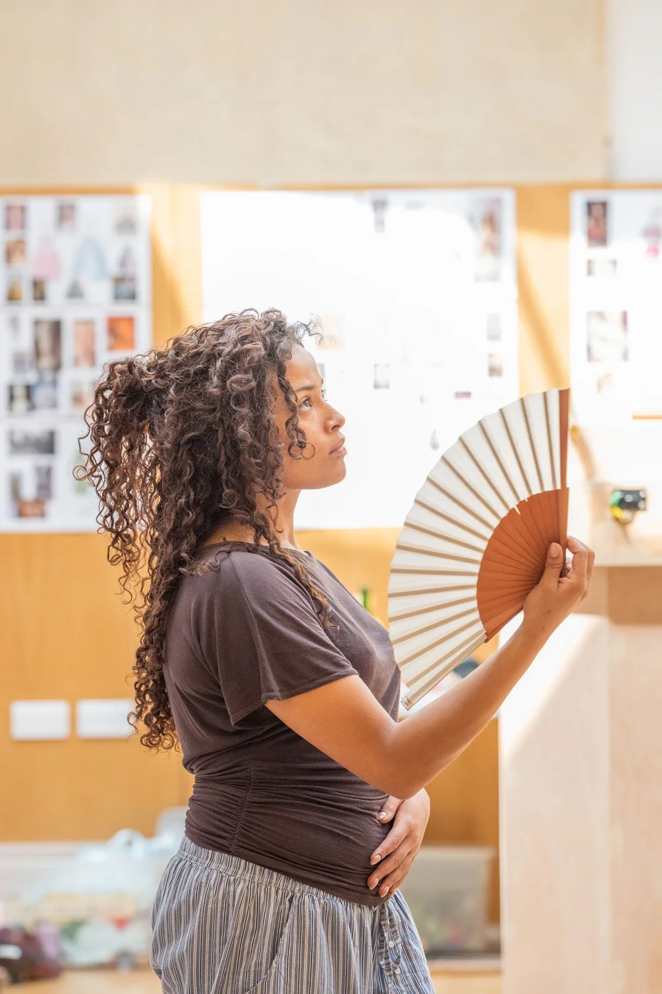 An average height, mixed race girl stands and holds an open fan in a brightly lit room.