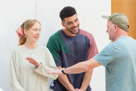 Three people stand together: a white, tall, ginger woman with freckles who wears a bandana and a long-sleeved striped t-shirt, a tall, mixed race British and Jamaican man wearing a blue, green and red t-shirt, and a white man wearing a green cap.