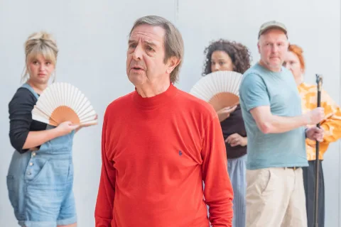 A person with brown hair with flecks of grey wearing a red top. Other people stand in the background, some holding fans.
