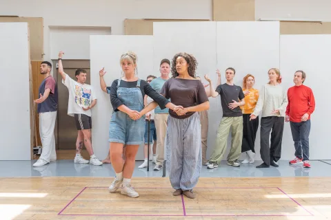 Two women, one white, with mousey brown hair and one mixed-race, stand with their arms interlinked. Behind them, a group of people stand leaning against a wall.
