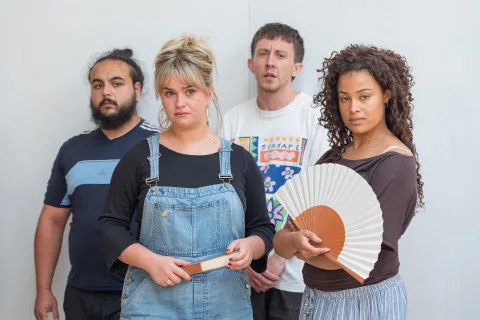Four people stand together in a group, with two of them standing in front of the other. One of the people at the front is a white mid-sized woman wearing dungarees with mousey-blonde hair, the other holds a fan and is an average height, mixed race girl.