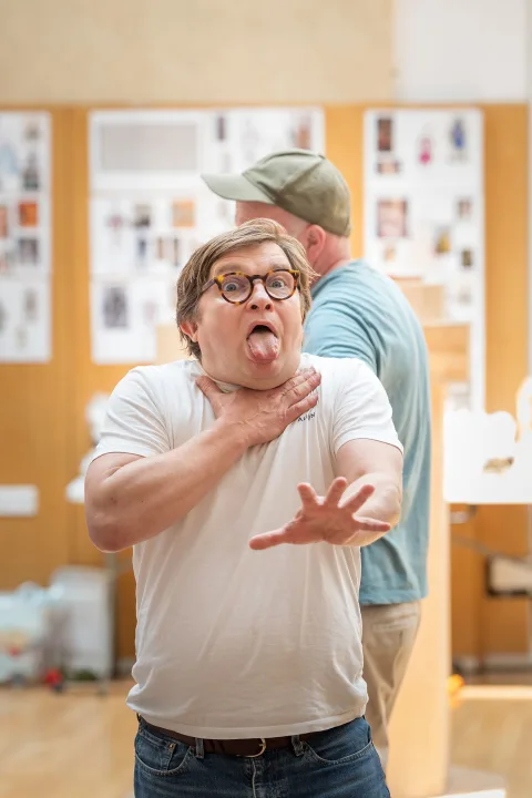 A white male with short blonde hair and glasses holds one hand to his throat and sticks out his tongue inside a brightly lit room.