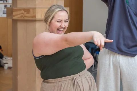 A white female, average height with medium blonde hair, stands smiling with one hand on her hip and one hand pointing in front of her.