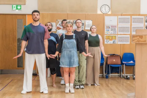 A group of people stand facing ahead, with their arms down by their sides and their hands pointing out. They are in a room with wooden walls and wooden floors.