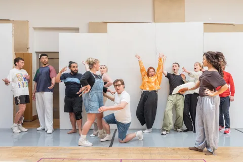 A diverse group of people in a wooden rehearsal room dance and throw their hands up in exclamation. Some hold fans, some kneel on the floor.