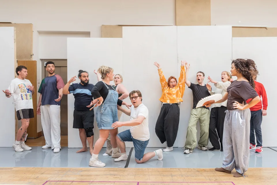 A diverse group of people in a wooden rehearsal room dance and throw their hands up in exclamation. Some hold fans, some kneel on the floor.