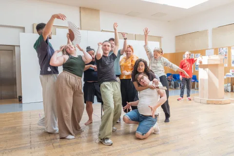 A diverse group of people raise their arms and dance in a wooden rehearsal room. One person holds a fan, and one person wearing a red top, leaning on a wooden pillar, watches from the side.
