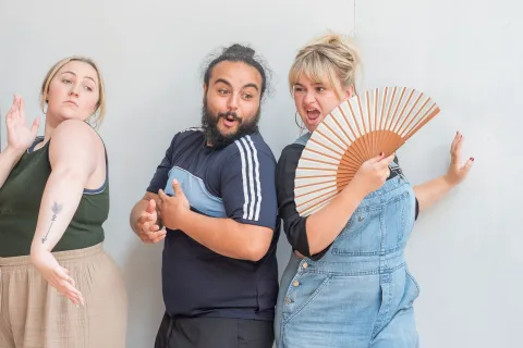 A white woman with blonde hair, wearing blue denim dungarees and holding a fan, stands next to a mixed Asian and British man with black hair and a bar. Next to them stands a white woman with blonde hair in a ponytail, wearing a green top.