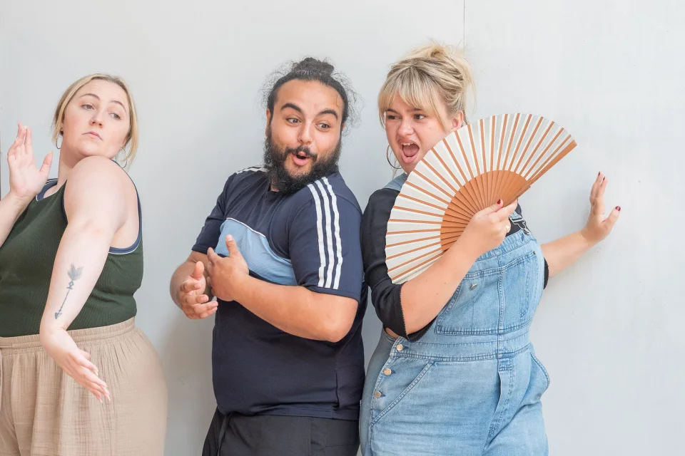 A white woman with blonde hair, wearing blue denim dungarees and holding a fan, stands next to a mixed Asian and British man with black hair and a bar. Next to them stands a white woman with blonde hair in a ponytail, wearing a green top.