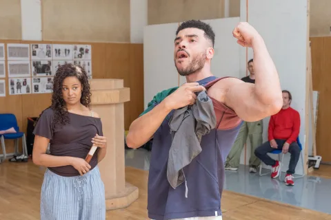 An mixed race girl stands and watches a mixed race British and Jamaican man pull back his t-shirt sleeve and flex his arm muscles.