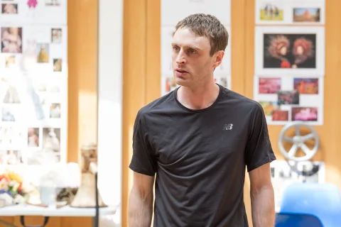 A white cis male with slim build and short brown hair, wearing a dark t-shirt, standing in front of a wall with coloured images stuck to it.
