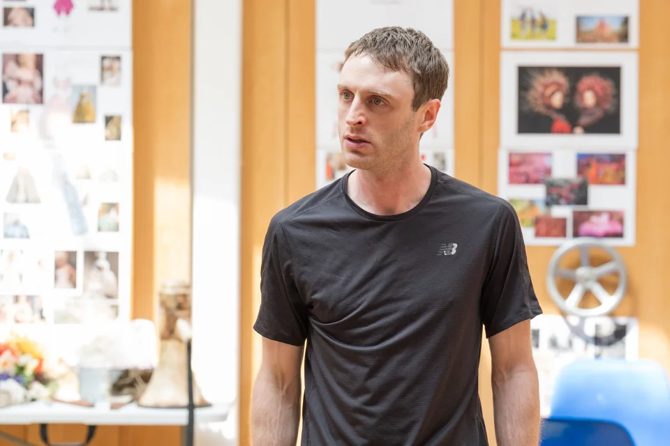A white cis male with slim build and short brown hair, wearing a dark t-shirt, standing in front of a wall with coloured images stuck to it.