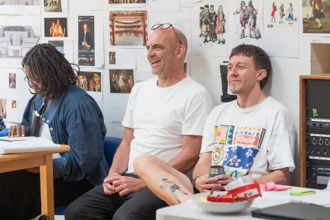 Three people sit against a white wall covered in pictures. One looks down at a script, whilst the other two smile at something in front of them.