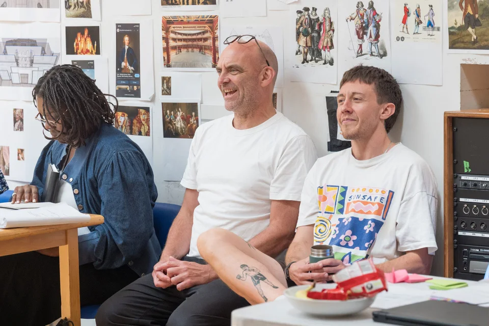 Three people sit against a white wall covered in pictures. One looks down at a script, whilst the other two smile at something in front of them.