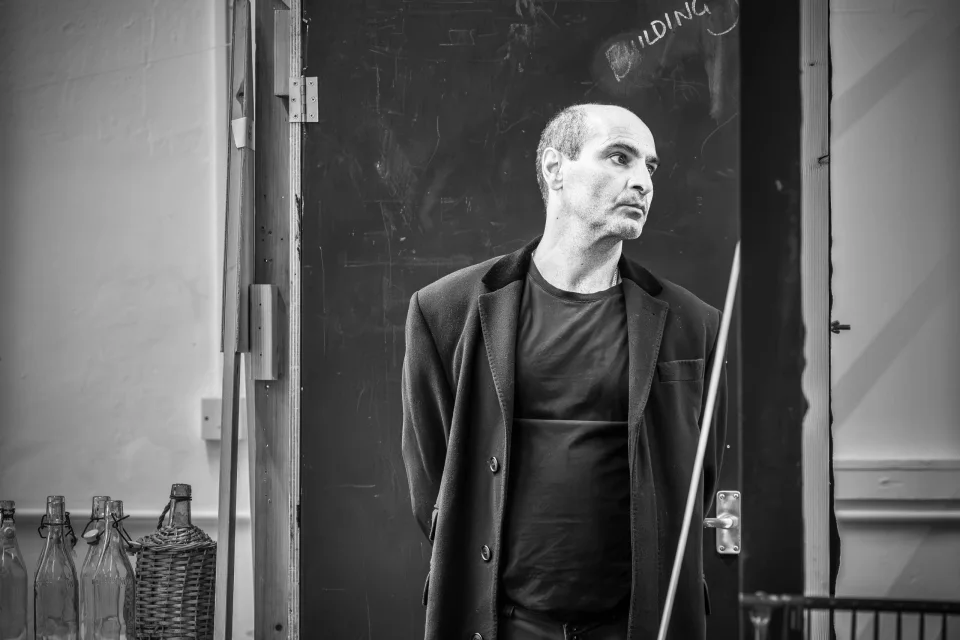 Zubin Varla stands in a long black coat with his hands behind his back. He looks to the left hand side with a focussed expression on his face.