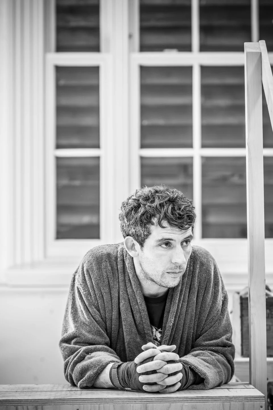 Josh O'Connor leans forwards on a step with his hands clasped. He wears a towelled robe and fingerless gloves.
